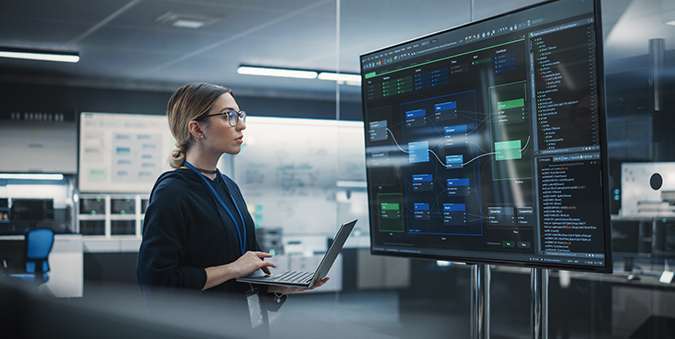 A woman in glasses holds a laptop while analyzing complex data on a large monitor. The setting is a modern, high-tech office. She appears focused and thoughtful.
